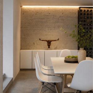 Comedor moderno con mesa blanca, sillas Eames, muro de piedra iluminado, cabeza de toro decorativa y cava de vinos. Diseño por MMS Arquitectos en Estado de México y Ciudad de México