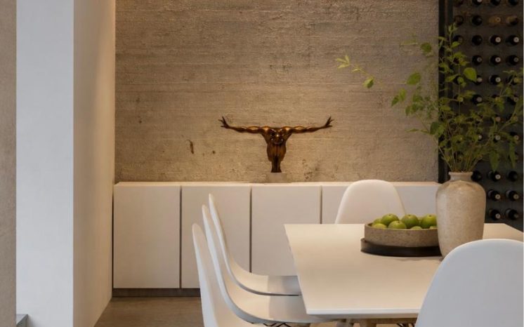 Comedor moderno con mesa blanca, sillas Eames, muro de piedra iluminado, cabeza de toro decorativa y cava de vinos. Diseño por MMS Arquitectos en Estado de México y Ciudad de México